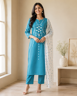 A woman modeling a teal blue three-piece salwar kameez suit with white embroidered details, matching blue pants, and a white dupatta with blue floral prints.