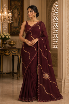 A woman models a deep maroon/burgundy saree with gold zari embroidery. The saree features a solid body, a draped pallu, and scalloped edges with embroidered details.