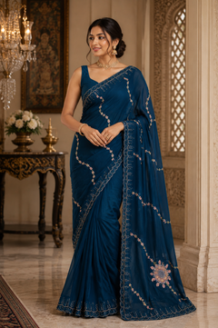 A woman wearing a deep teal blue saree with silver and gold embroidery, standing in an ornate, traditional Indian room with a chandelier and carved walls.