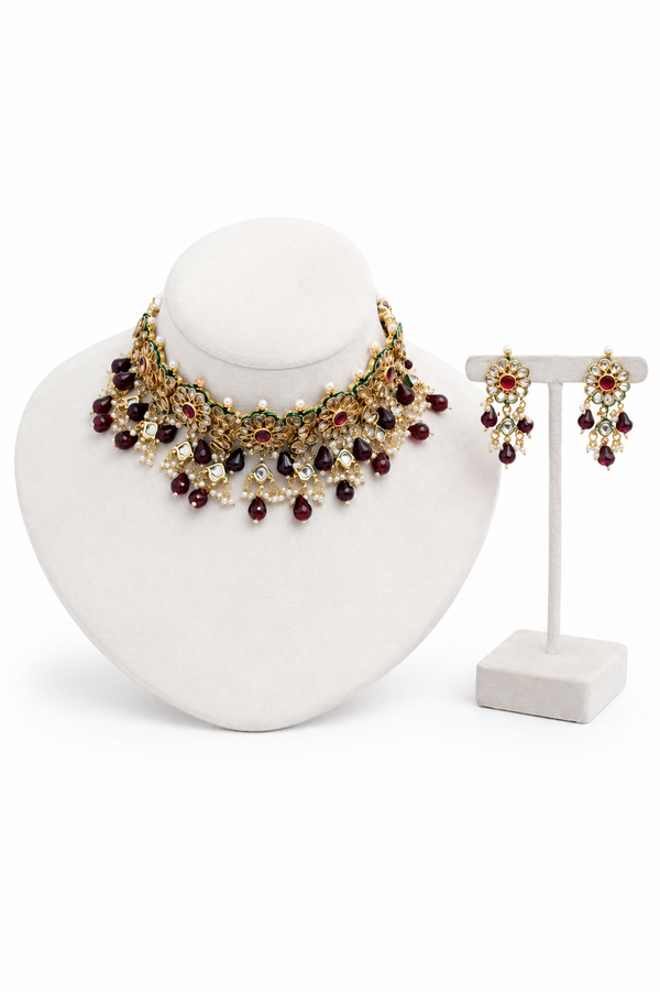 An ornate gold-toned statement necklace displayed on a white jewelry bust, with matching drop earrings. The jewelry features dark red beads, clear rhinestones, and green leaf-like accents.