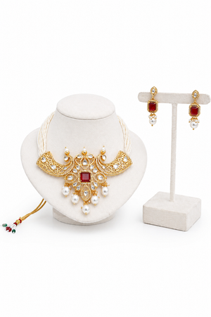 Gold-toned traditional jewelry set featuring a statement necklace and matching drop earrings on a stand, with white pearls, red stones, and small multicolor bead accents.