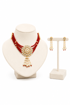 A red beaded necklace with a gold-toned, pearl-embellished circular pendant is displayed on a cream jewelry bust, alongside matching gold and pearl dangle earrings on a small stand against a white background.