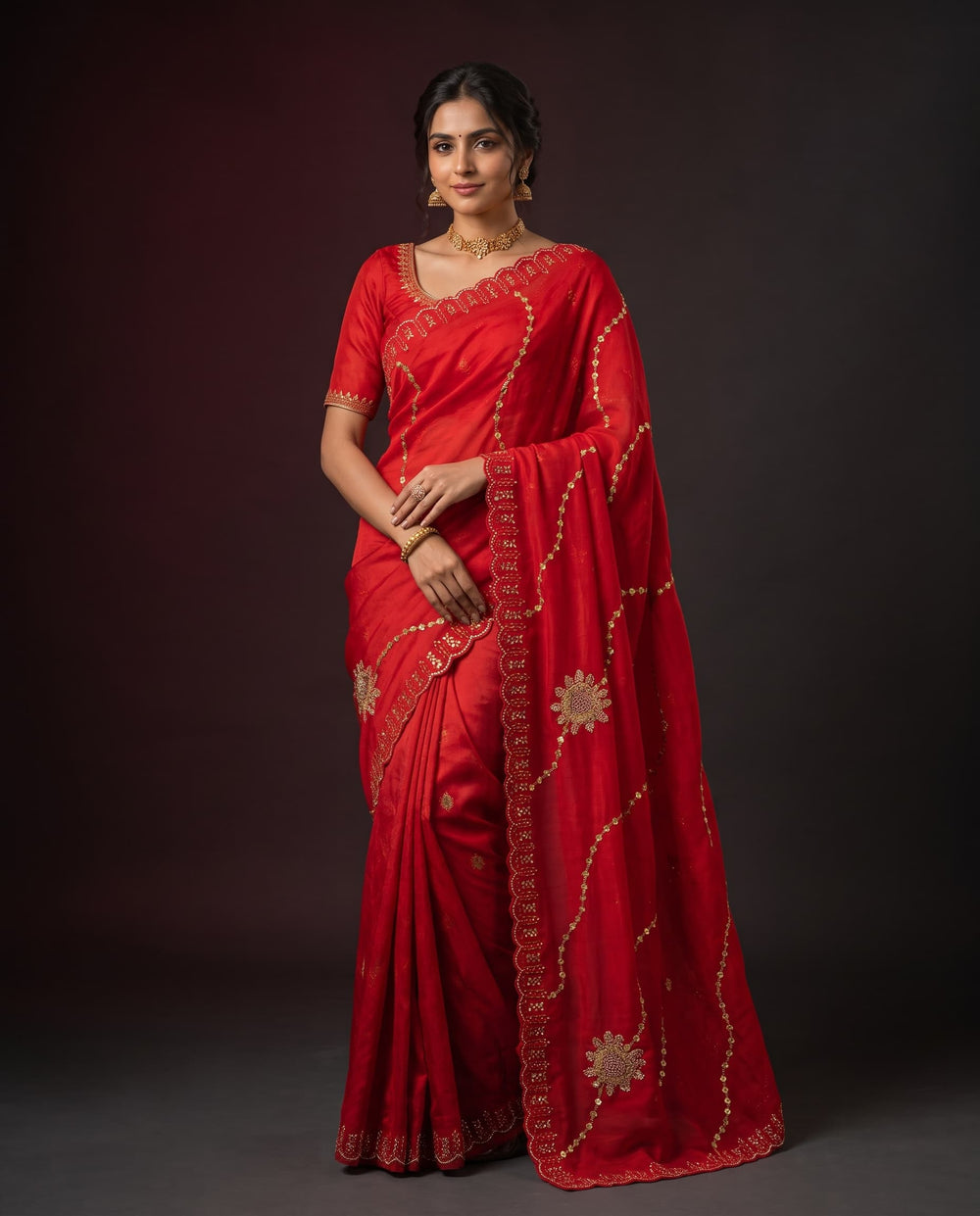 A woman modeling a bright red saree with gold floral and wavy embroidered details, paired with a matching red blouse, against a dark studio background.