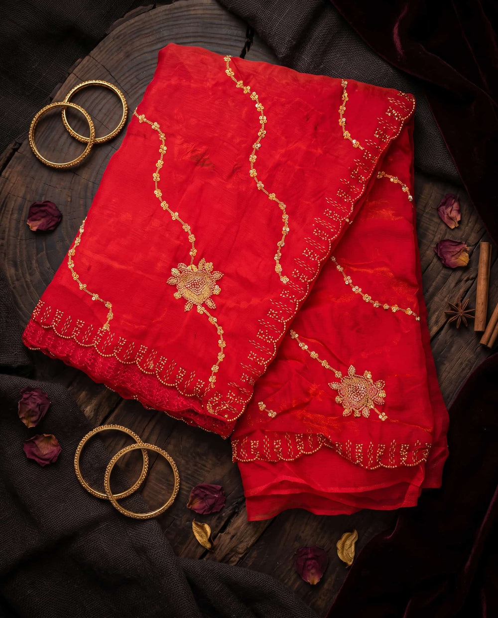A woman modeling a bright red saree with gold floral and wavy embroidered details, paired with a matching red blouse, against a dark studio background.