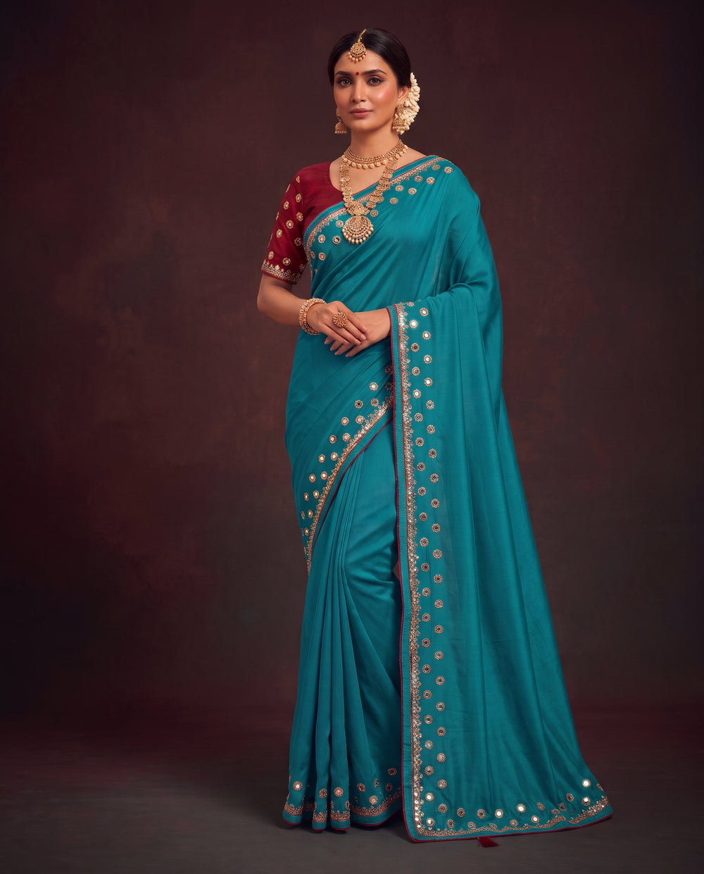 A woman modeling a teal blue saree with gold embroidered borders and a deep red blouse, standing against a dark brown studio backdrop.