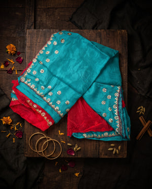 A folded turquoise and red saree with ornate gold embroidered borders and small floral motifs, displayed on a dark wooden board with dried flowers, bangles, and spices around it.