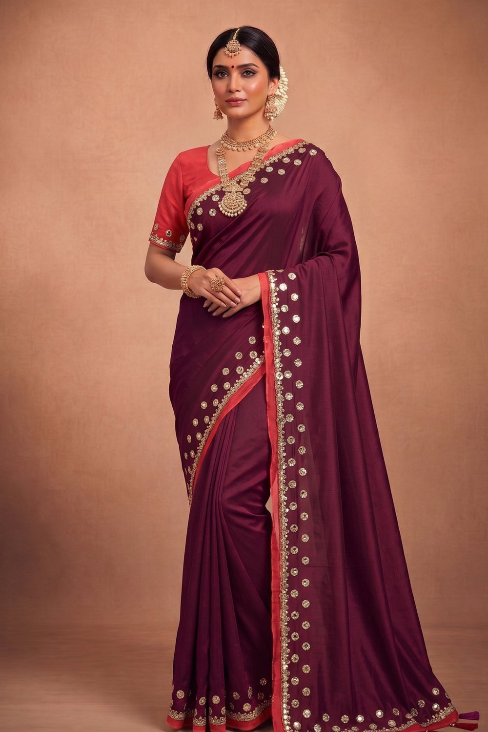 A woman models a deep maroon purple saree with a red blouse, decorated with gold mirror work and a red border, set against a solid brown studio backdrop.