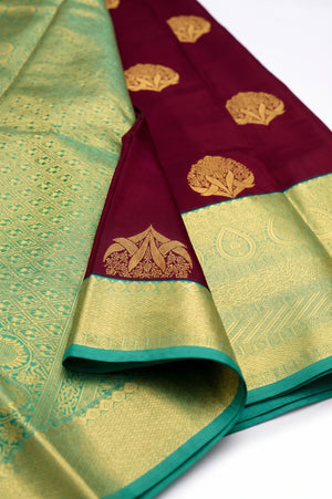 A close-up of a folded silk saree with a deep maroon border, gold zari floral motifs, and a contrasting light green body with intricate gold woven patterns.