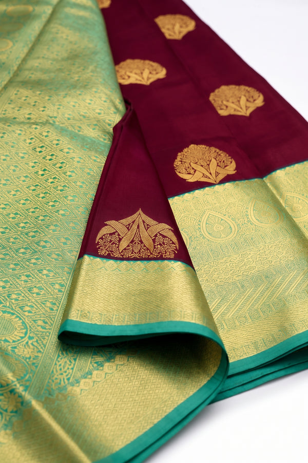 A close-up of a folded silk saree with a deep maroon border, gold zari floral motifs, and a contrasting light green body with intricate gold woven patterns.