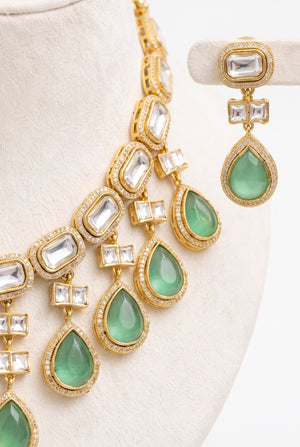 Gold-toned statement jewelry set featuring a layered necklace and matching drop earrings with green teardrop gemstones and clear rectangular stones.