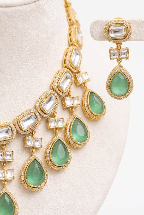 Gold-toned statement jewelry set featuring a layered necklace and matching drop earrings with green teardrop gemstones and clear rectangular stones.