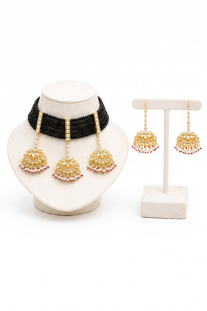 A traditional Indian jewelry set featuring a black beaded collar necklace with three gold bell-shaped dangle earrings adorned with red and white beads, displayed on a cream-colored jewelry stand against a white background.