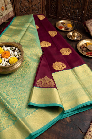 A folded silk saree, featuring a deep maroon body with gold zari motifs, a light green border, and a teal edge. 