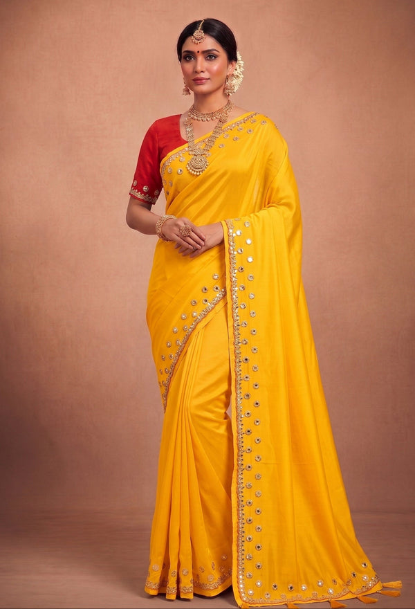 A woman modeling a bright yellow saree with a red sleeve and silver mirror work along the border, posed against a solid brown studio backdrop.