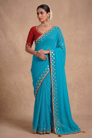A woman modeling a turquoise blue saree with a red sleeve blouse, decorated with gold and silver mirror work borders and floral embroidery, set against a textured beige studio backdrop.
