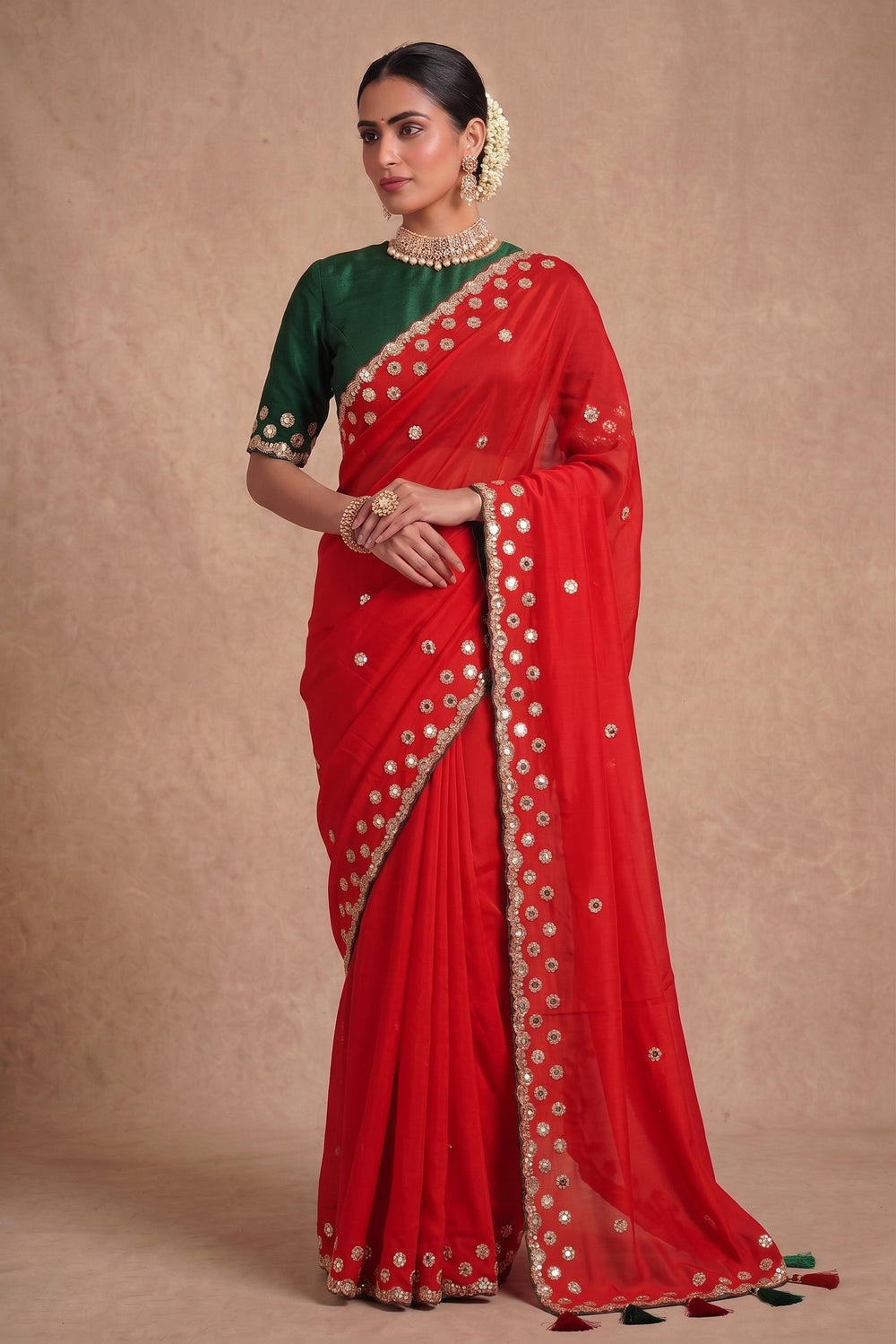 A woman modeling a red georgette saree with a dark green blouse, decorated with gold zari mirror work and embroidered borders.