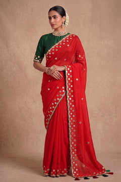 A woman modeling a red georgette saree with a dark green blouse, decorated with gold zari mirror work and embroidered borders.