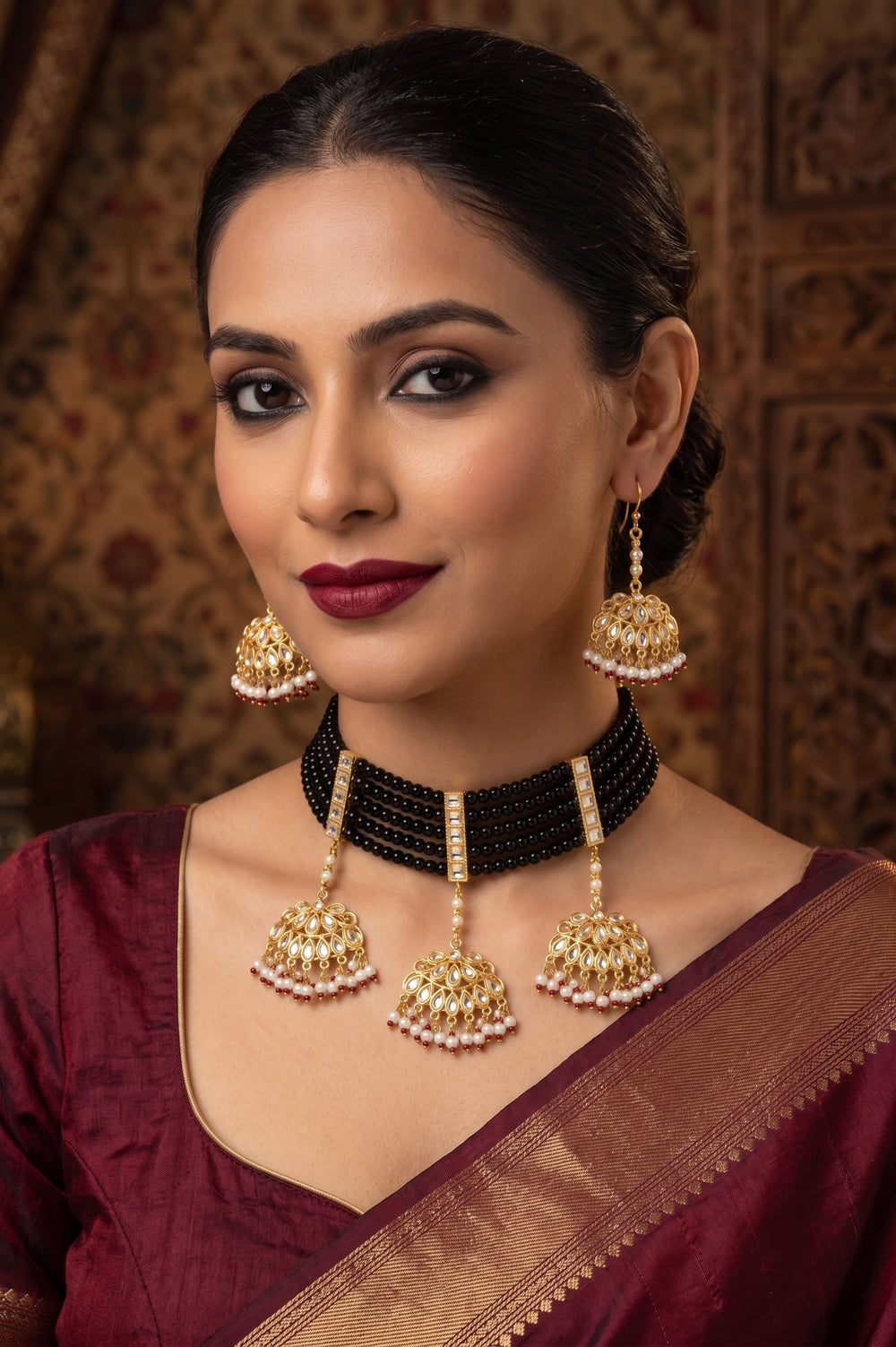 A traditional Indian jewelry set featuring a black beaded collar necklace with three gold bell-shaped dangle earrings adorned with red and white beads, displayed on a cream-colored jewelry stand against a white background.