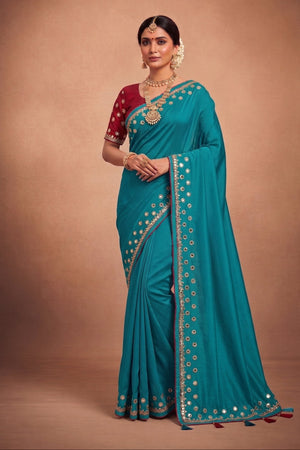 A woman modeling a teal blue saree with a deep red sleeve and gold mirror work borders, posed against a solid brown studio backdrop.