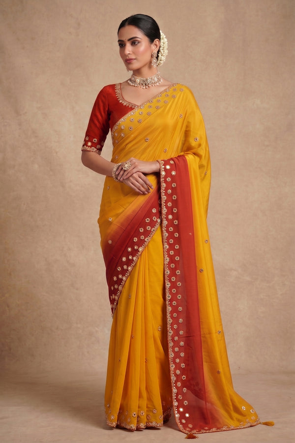 A woman modeling a yellow and red saree with gold mirror work and embroidery, standing against a textured beige studio backdrop.