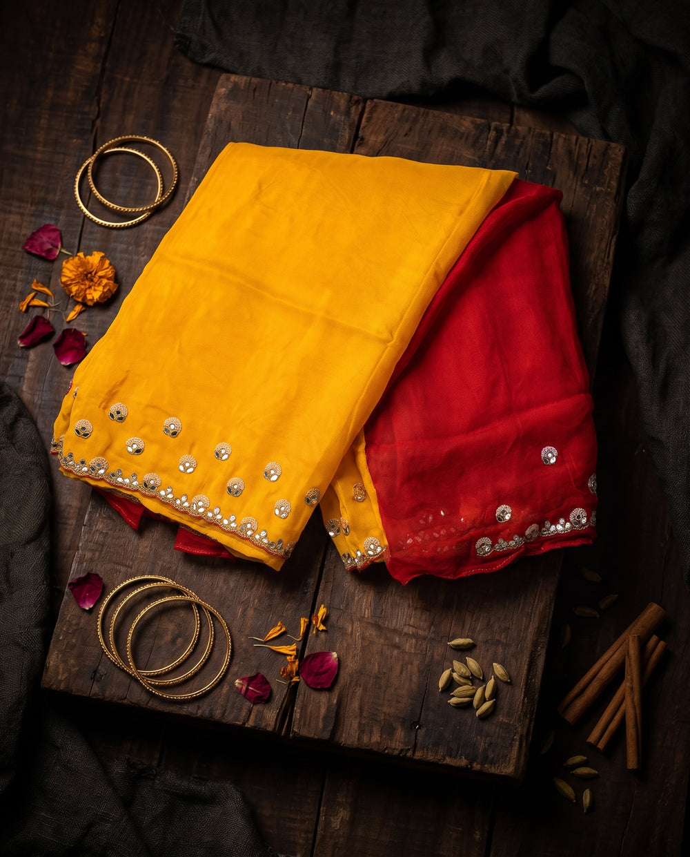 A woman modeling a bright yellow saree with a red sleeveless blouse, decorated with gold mirror work along the border and pallu, set against a dark brown studio background.