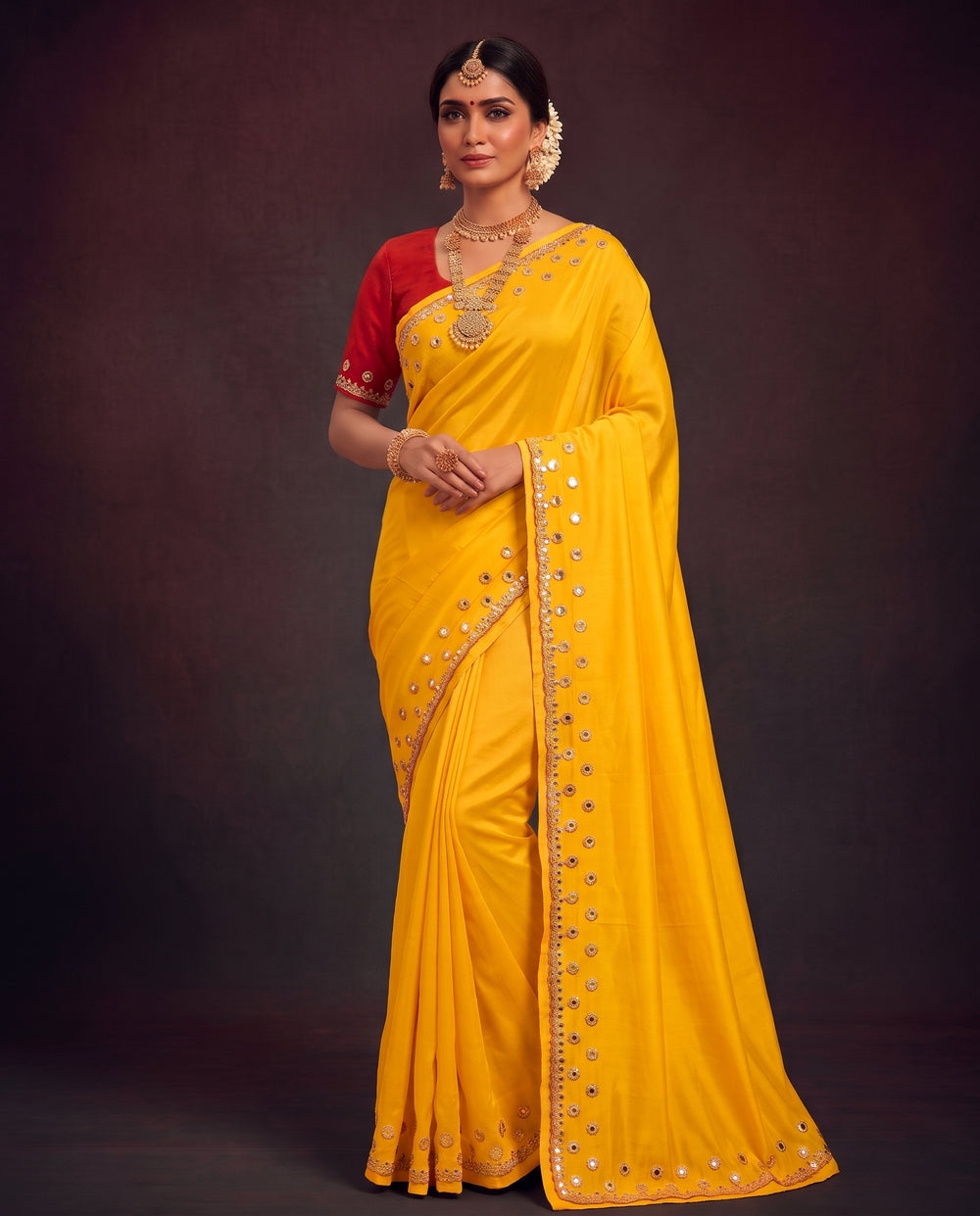 A woman modeling a bright yellow saree with a red sleeveless blouse, decorated with gold mirror work along the border and pallu, set against a dark brown studio background.