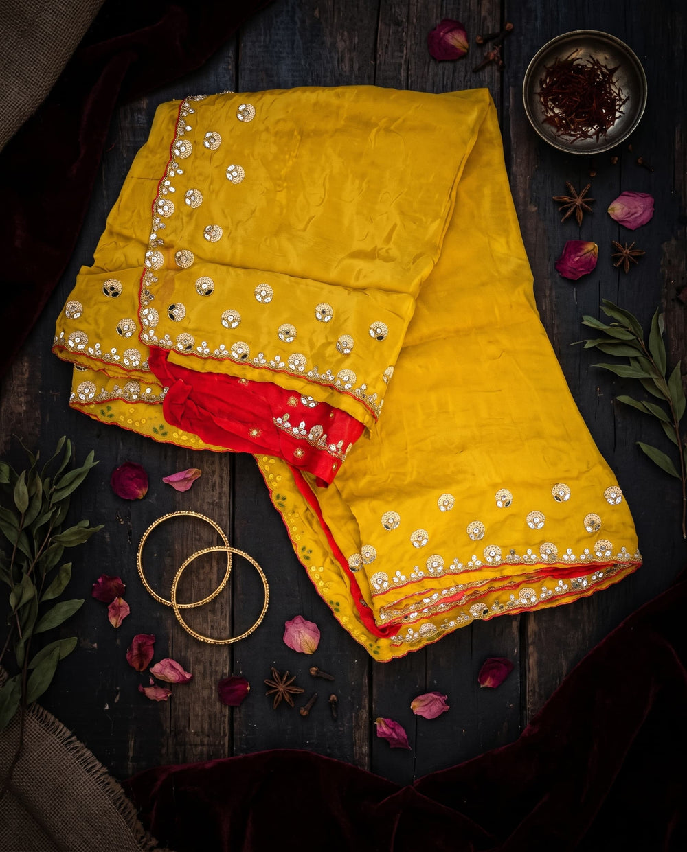 A woman modeling a bright yellow saree with a red sleeveless blouse, decorated with gold mirror work and embroidery along the border, set against a dark brown studio background.