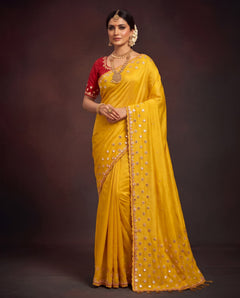 A woman modeling a bright yellow saree with a red sleeveless blouse, decorated with gold mirror work and embroidery along the border, set against a dark brown studio background.