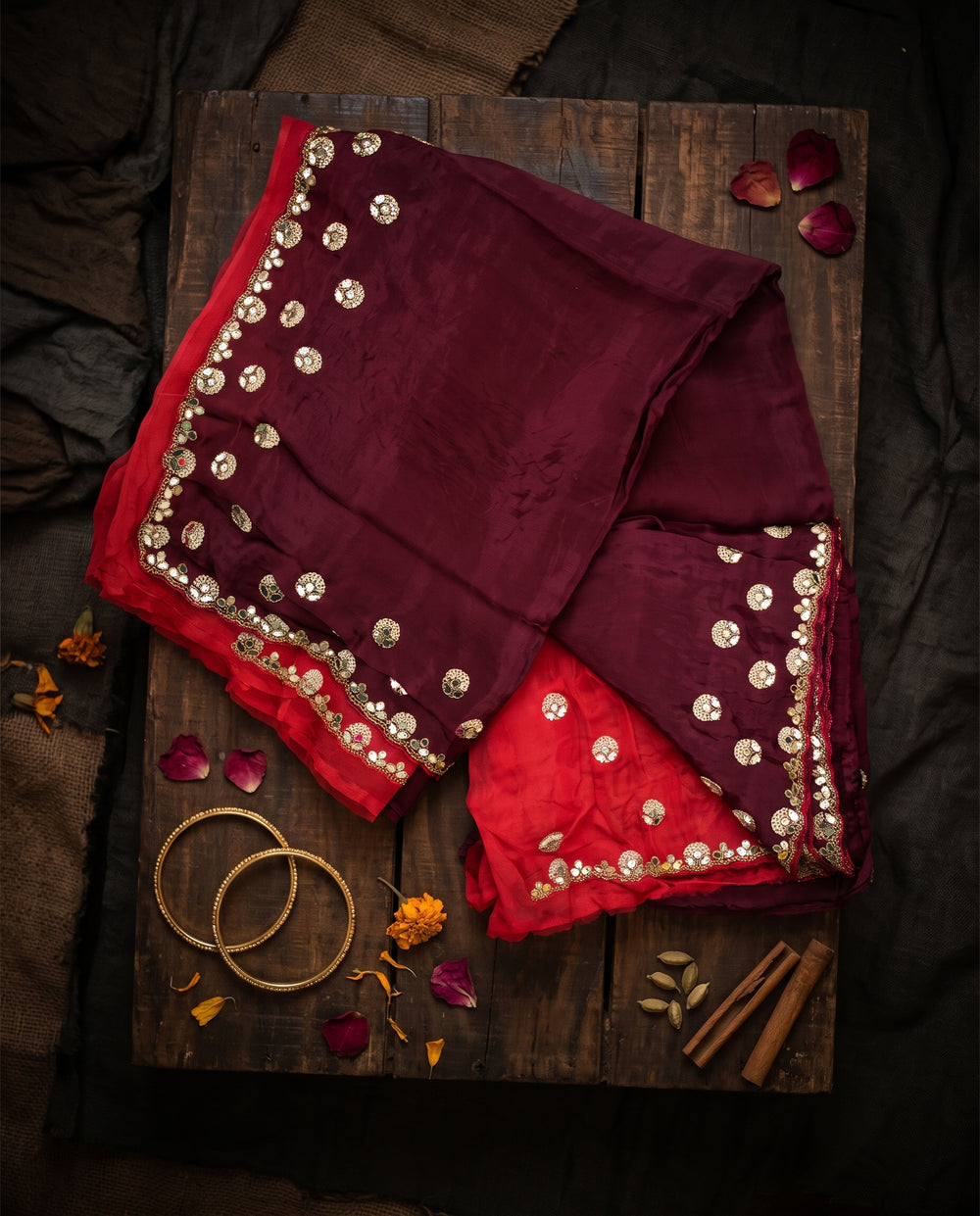 A woman modeling a deep maroon saree with a red-orange blouse and gold mirror work border, set against a dark brown studio backdrop.