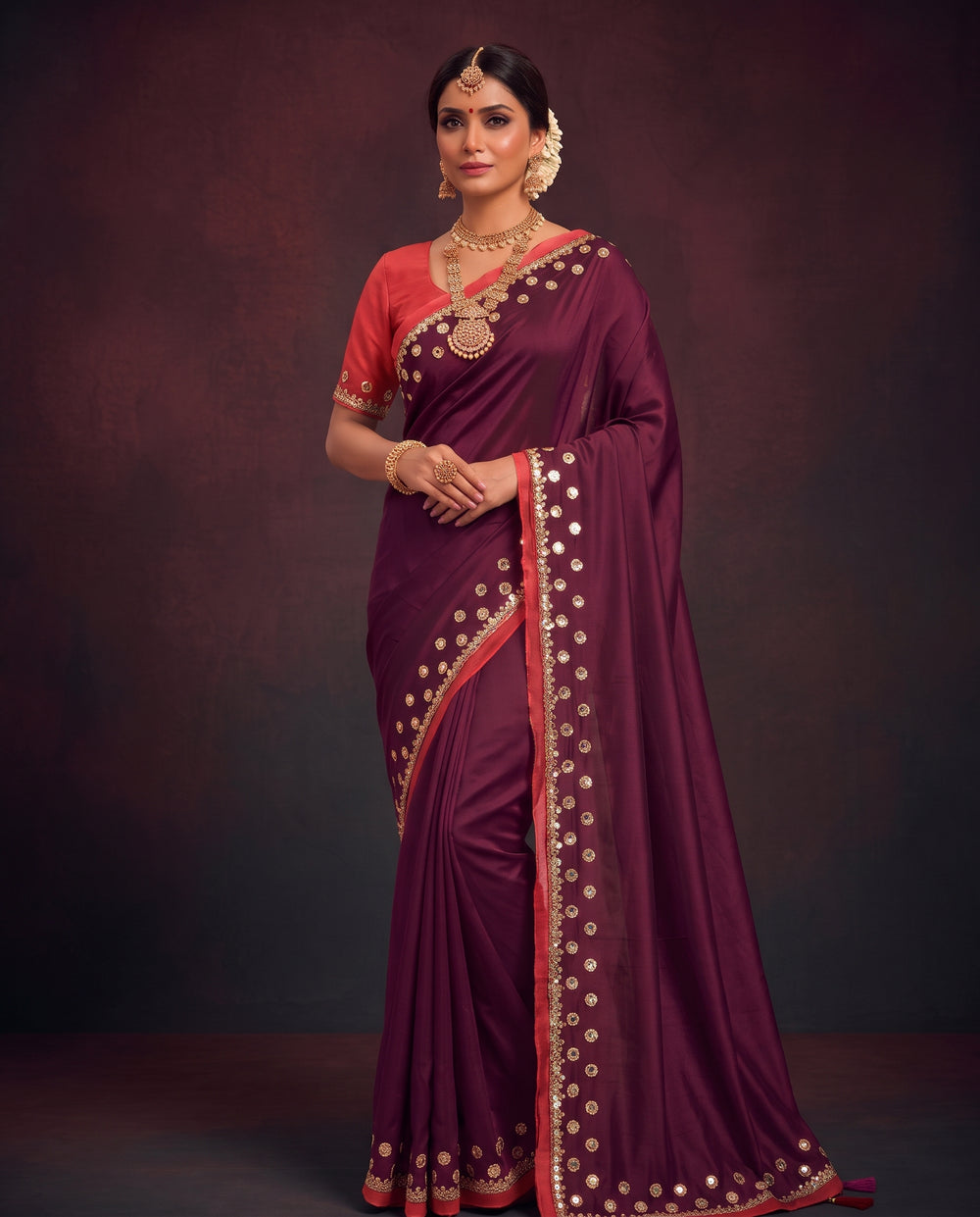 A woman modeling a deep maroon saree with a red-orange blouse and gold mirror work border, set against a dark brown studio backdrop.