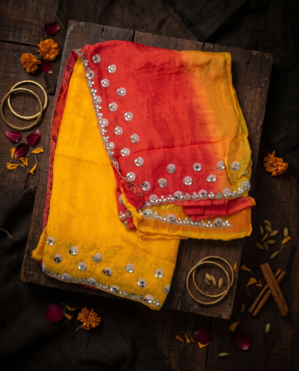 A woman modeling a traditional Indian saree with a mustard yellow body, a red border, and a red sleeve, set against a dark brown studio background.