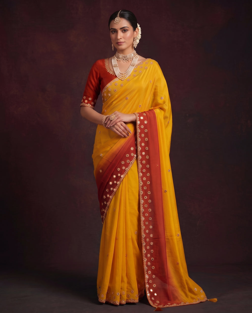 A woman modeling a traditional Indian saree with a mustard yellow body, a red border, and a red sleeve, set against a dark brown studio background.