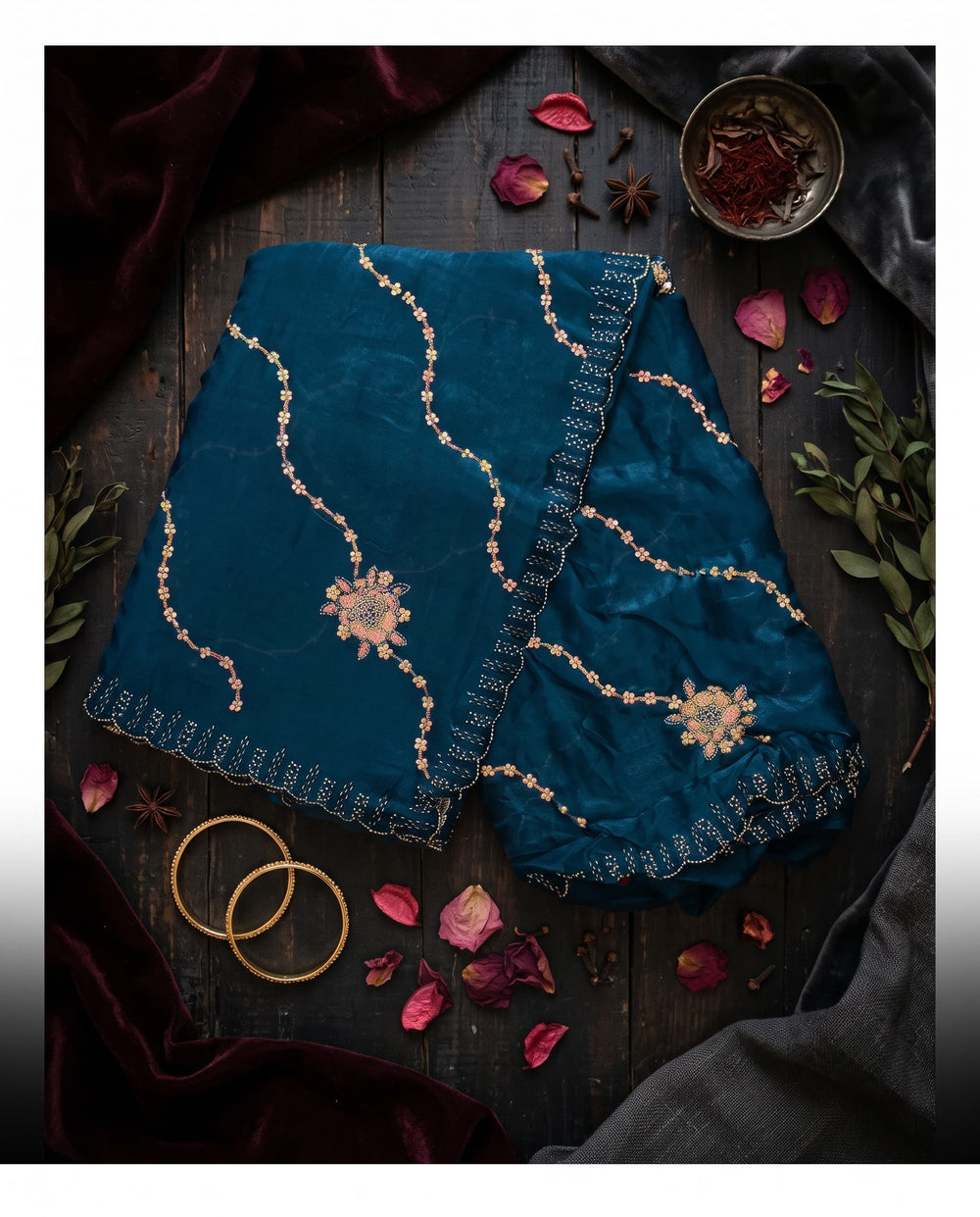 A woman modeling a deep teal blue saree with silver embroidered patterns and a matching border, set against a dark brown studio backdrop.