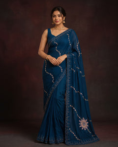 A woman modeling a deep teal blue saree with silver embroidered patterns and a matching border, set against a dark brown studio backdrop.