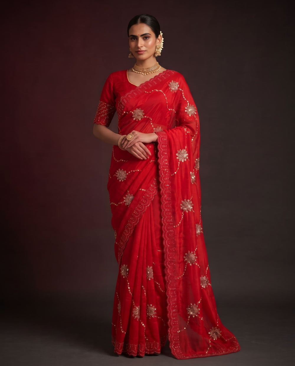 A woman modeling a bright red saree with gold floral embroidery and a matching border, posed against a dark brown studio background.