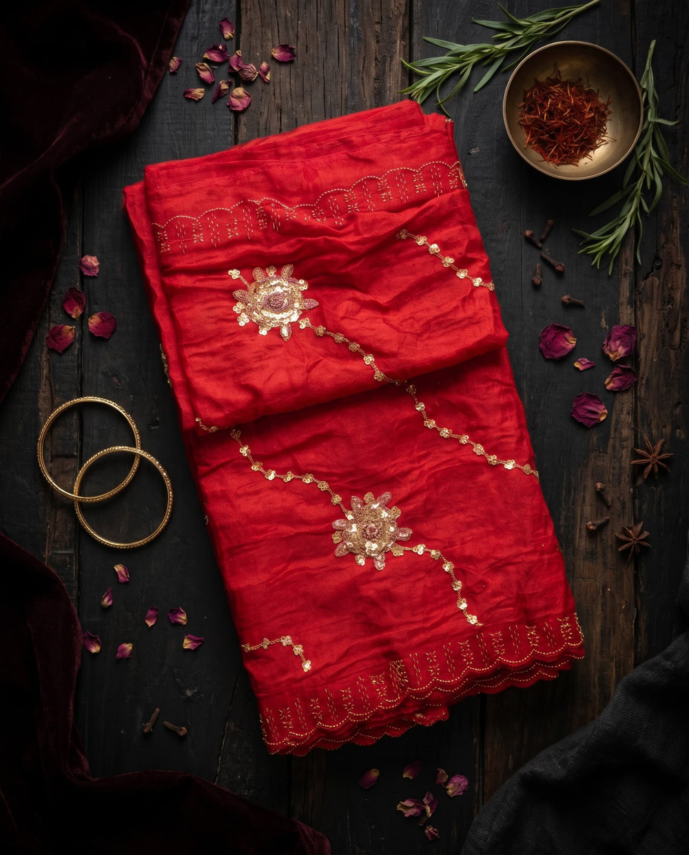 A woman modeling a bright red saree with gold floral embroidery and a matching border, posed against a dark brown studio background.