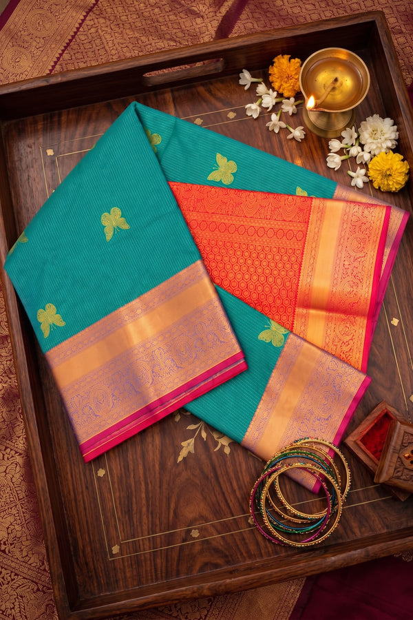 A folded teal and orange silk saree with gold zari borders and motifs, displayed on a dark wooden tray alongside a brass diya, white flowers, and stacked bangles.